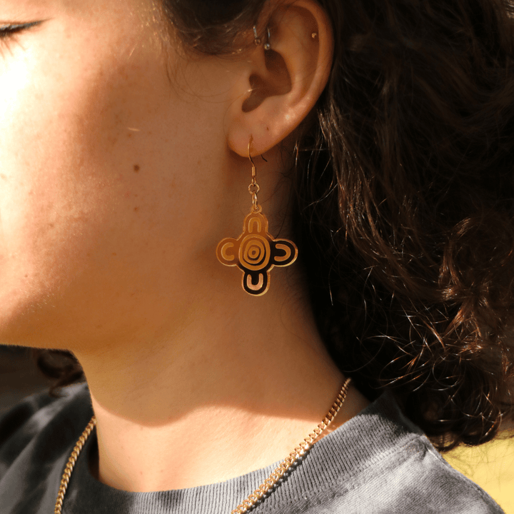 close up of model wearing haus of dizzy meeting place mirror earrings size small with a blurred natural background. the earring features aboriginal art which represents people coming together, seated at a specific site, camp or gathering place.