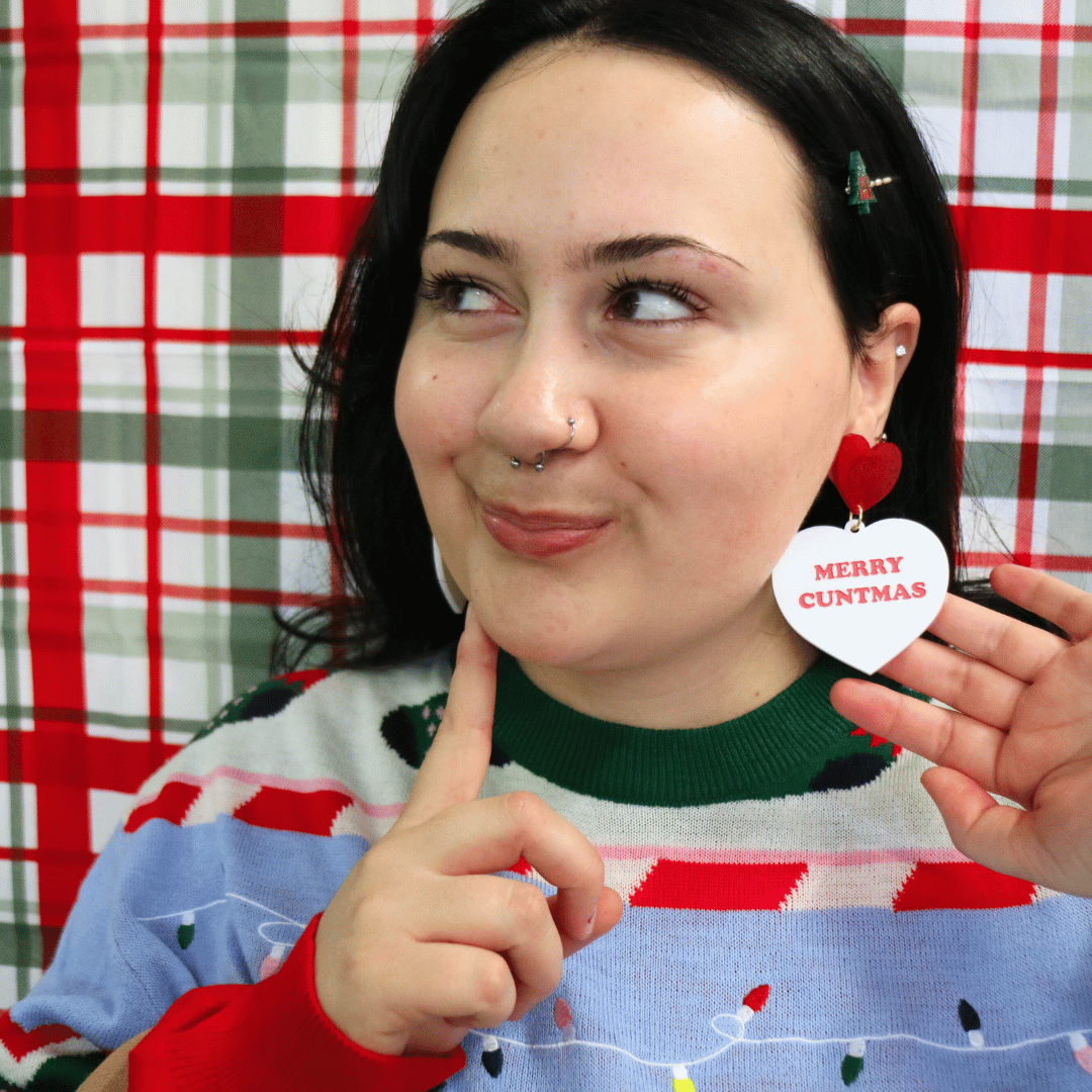 model wearing ugly christmas sweater with haus of dizzy merry cuntmas large earrings with red text, looking away from camera with cheeky face