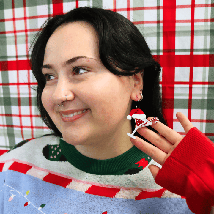 model wearing ugly christmas sweater against a red and green plaid background. she is wearing haus of dizzy merry martini earrings and looking away from camera