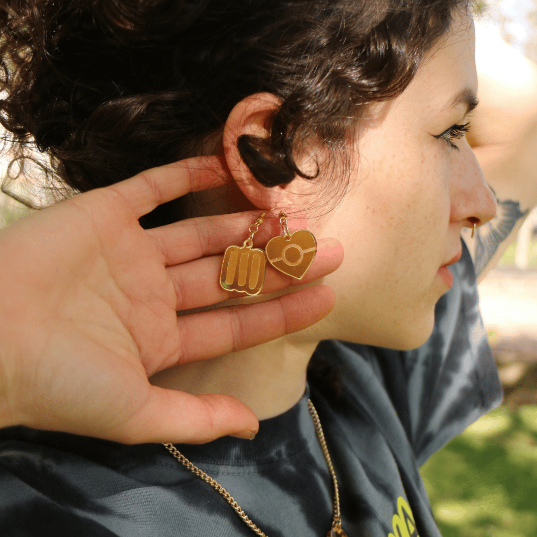 close up of model wearing haus of dizzy mini aboriginal flag mirror heart earrings and blak collection markings earrings both in gold mirror, she has stacked them in her ear piercings and is holding them up to the camera