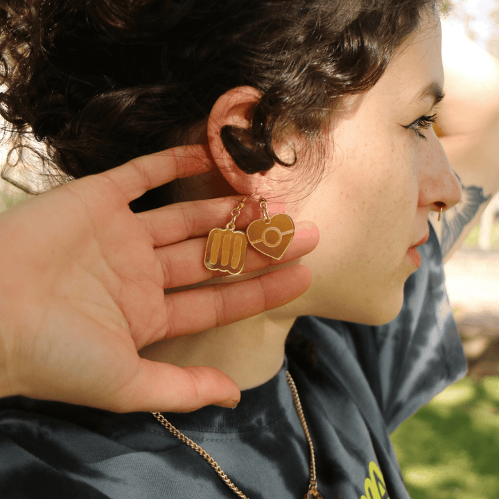 close up of model wearing haus of dizzy mini aboriginal flag mirror heart earrings and blak collection markings earrings both in gold mirror, she has stacked them in her ear piercings and is holding them up to the camera