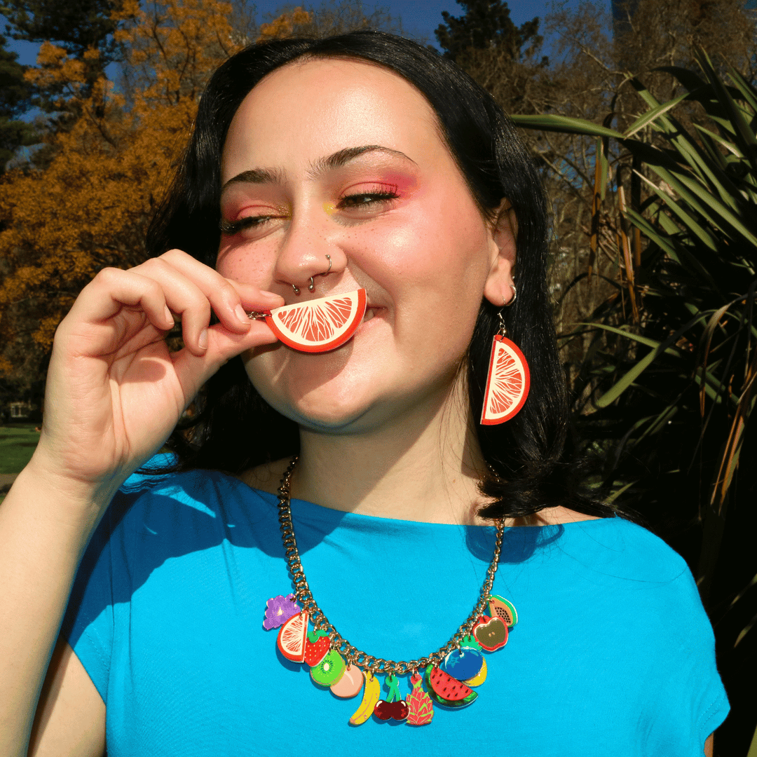 Smiling woman in a blue shirt wearing haus of dizzys vitamin c orange slice shaped earrings in one ear, holding the other up in front of her mouth smiling.