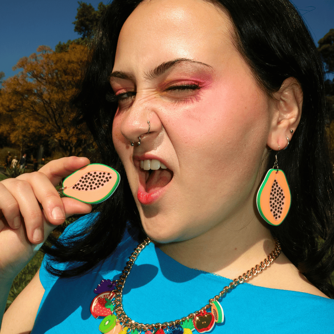 Close-up of a woman outdoors making a playful grimace, holding haus of dizzys papaya earrings with bold pink makeup