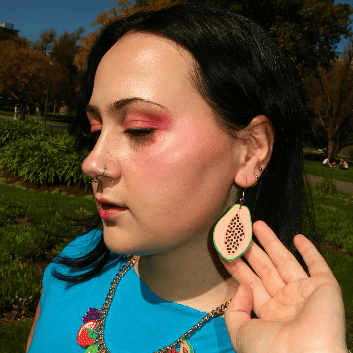 Close-up outdoor portrait of a woman with black hair, pink eye makeup, nose rings, and haus of dizzys papaya earrings held up in a sunny park.