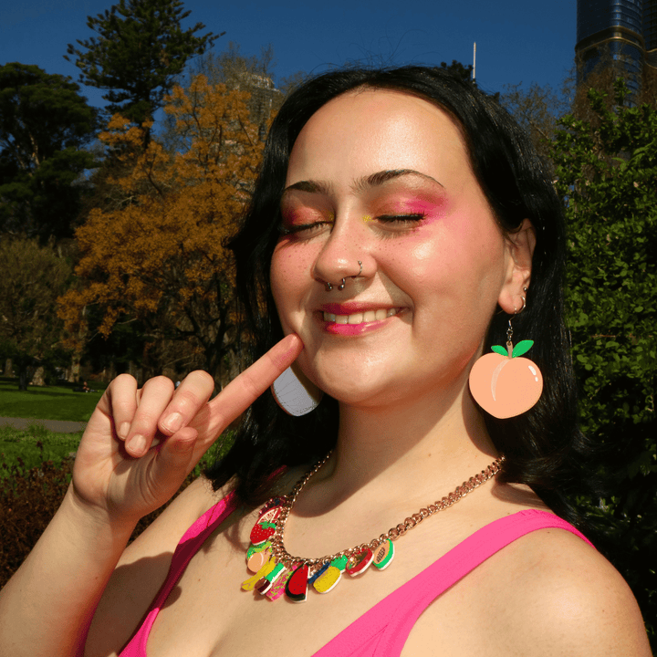 A smiling woman outdoors wearing haus of dizzy peachy keen peach-shaped dangling earrings and a colorful fruit-themed necklace, with a pink top and a park background.
