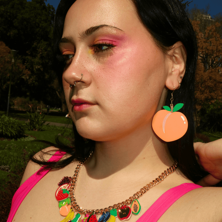 Close-up portrait of a woman with pink eyeshadow, nose ring, wearing haus of dizzys peach shaped dangle earrings, posing confidently outdoors in warm sunlight.