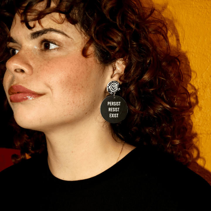 close up image of Aboriginal model wearing haus of dizzys mob only persist resist exist earrings. they feature a black circle with bold white text and a stud top which has aboriginal artwork on it.