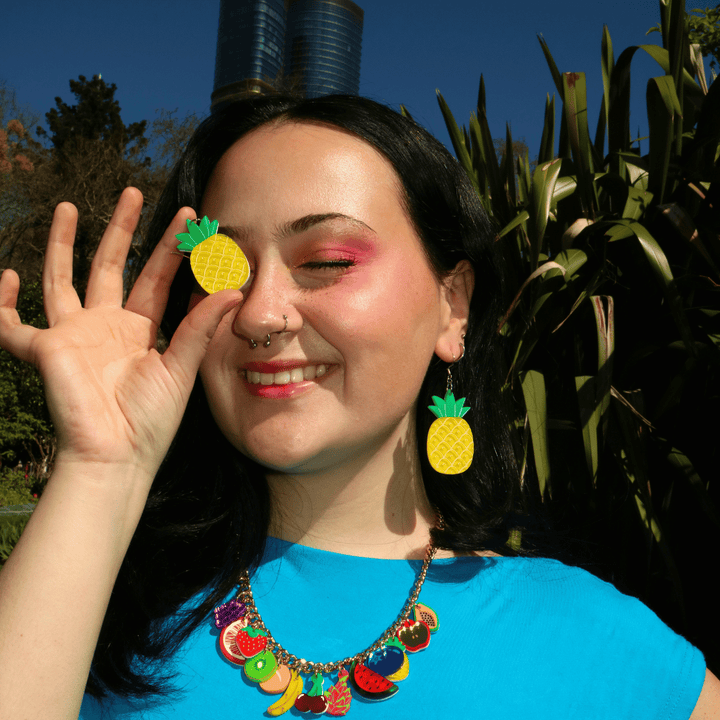 A woman outdoors wearing haus of dizzys bright yellow pineapple-shaped earrings and a colorful fruit charm necklace, holding a matching pineapple earring over one of her eyes.
