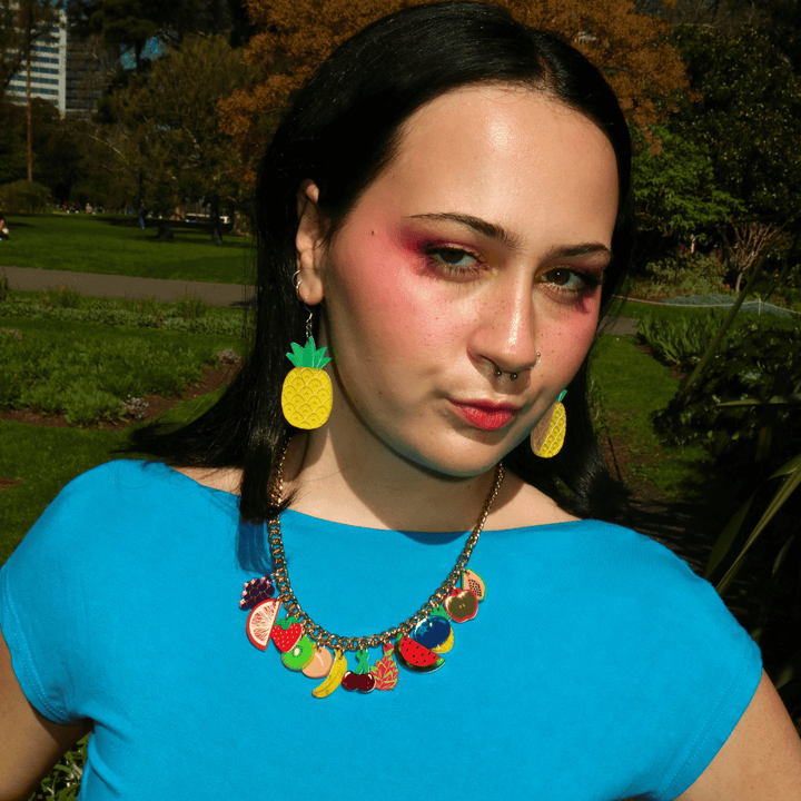 A woman outdoors wearing a bright blue top, a colorful fruit-themed charm necklace, and yellow pineapple-shaped earrings, with a park background.