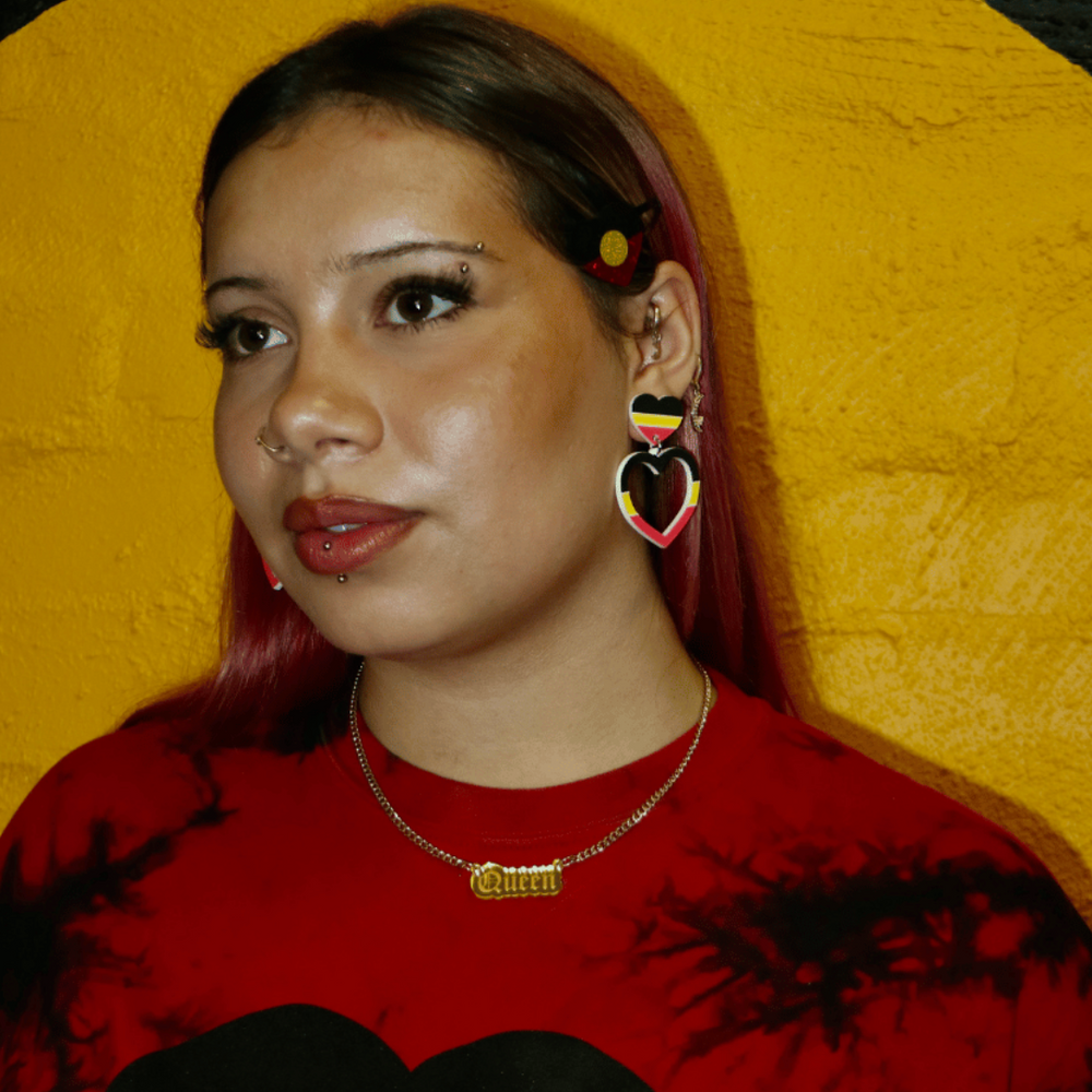 model wearing a red tie dye shirt with an Aboriginal flag design against a yellow and black background. she is wearing haus of dizzy queen plate necklace in small size in gold.