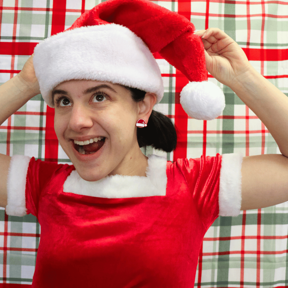 model wearing santa hat and dress with haus of dizzys santas swag studs in red gloss, looking away from camera playing with santa hat