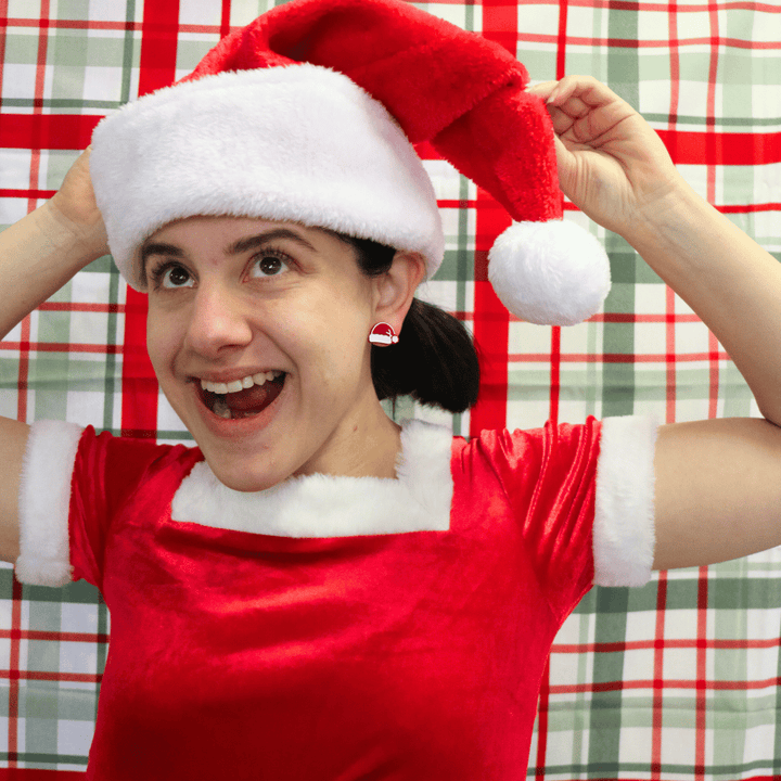 model wearing santa hat and dress with haus of dizzys santas swag studs in red gloss, looking away from camera playing with santa hat