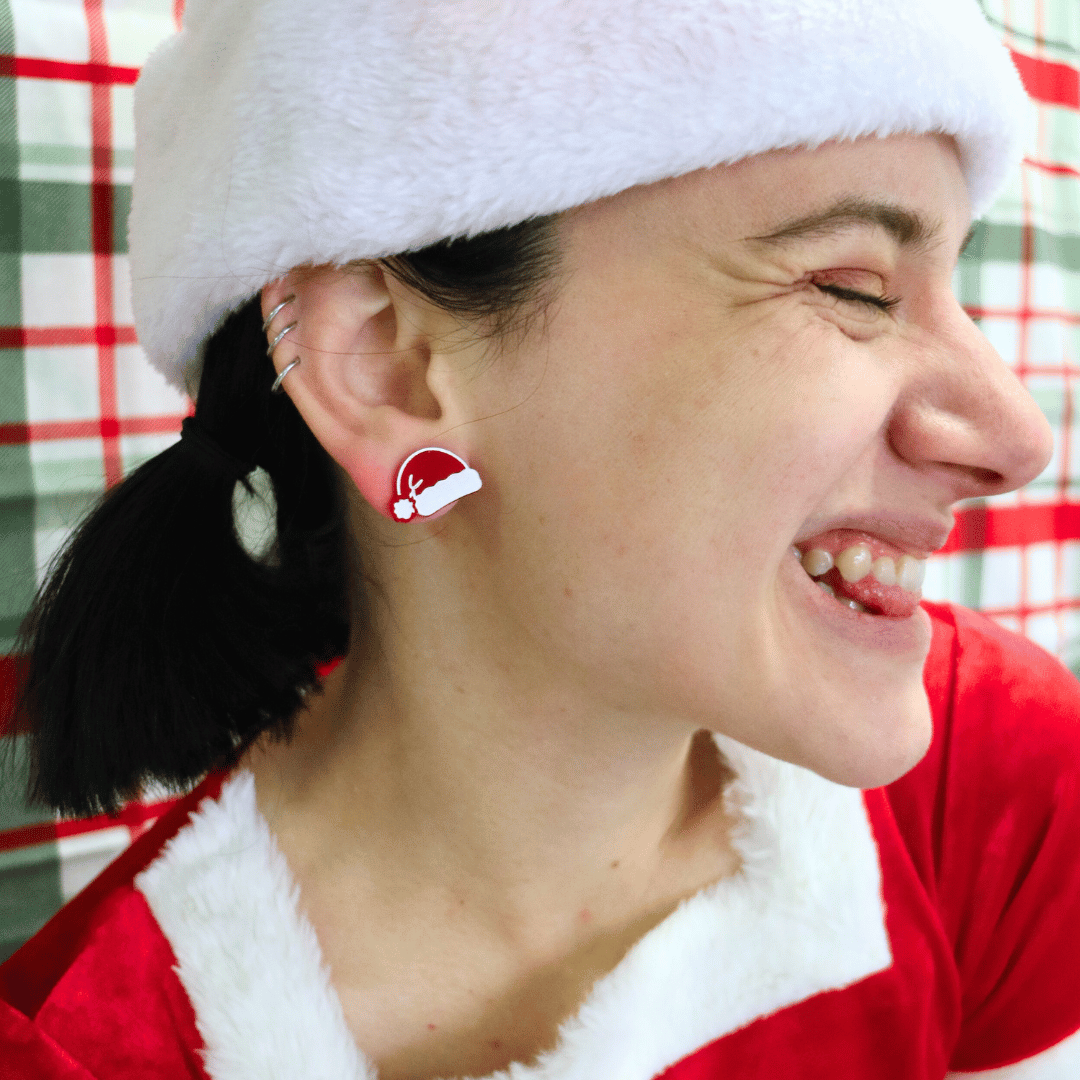 model wearing santa hat withhaus of dizzys santas swag studs in red gloss, looking away from camera making a cheeky face