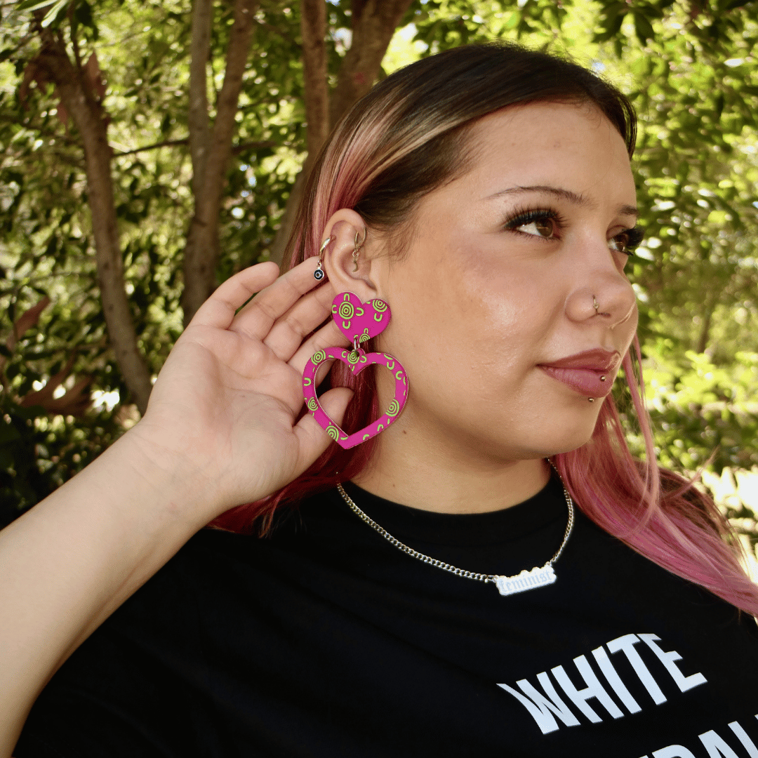 image of model in nature wearing haus of dizzy meeting place sea glass heart hoops in flamingo and apple green colour. 