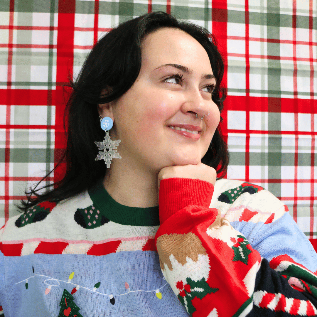 model wearing ugly christmas sweater with haus of dizzy snow hustler earrings in white glitter with head resting on fist looking away from camera.