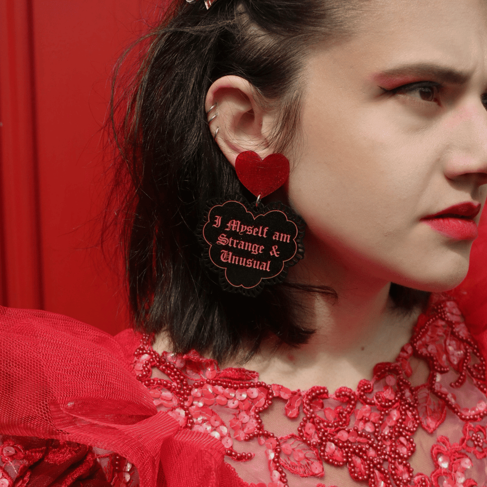 close-up of a model wearing haus of dizzy strange and unusual black and red glitter heart lace earrings with text that reads 'i myself am strange and unusual', against a red background
