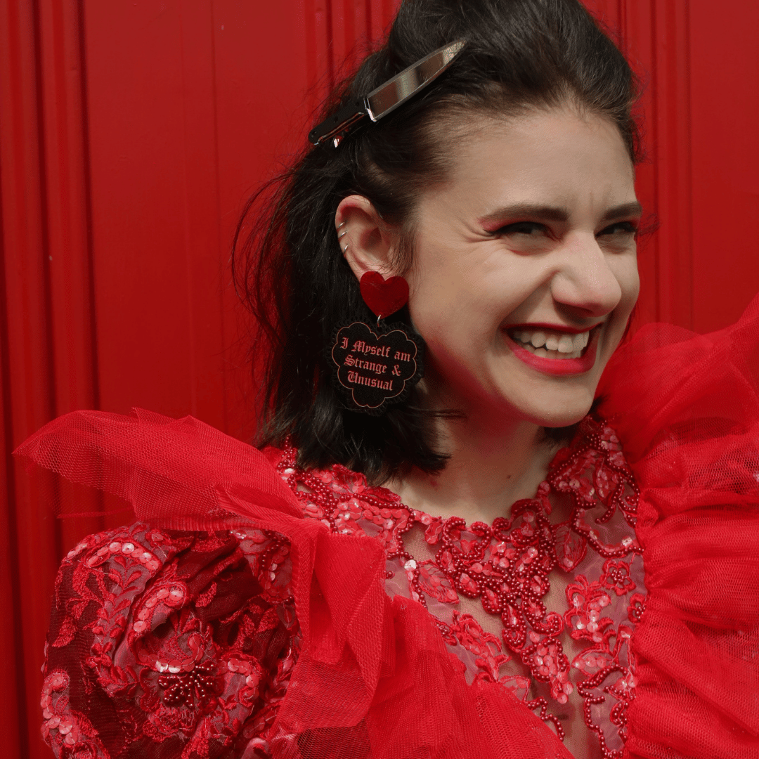 a model wearing haus of dizzy strange and unusual black and red glitter heart lace earrings with text that reads 'i myself am strange and unusual', against a red background, she is smiling