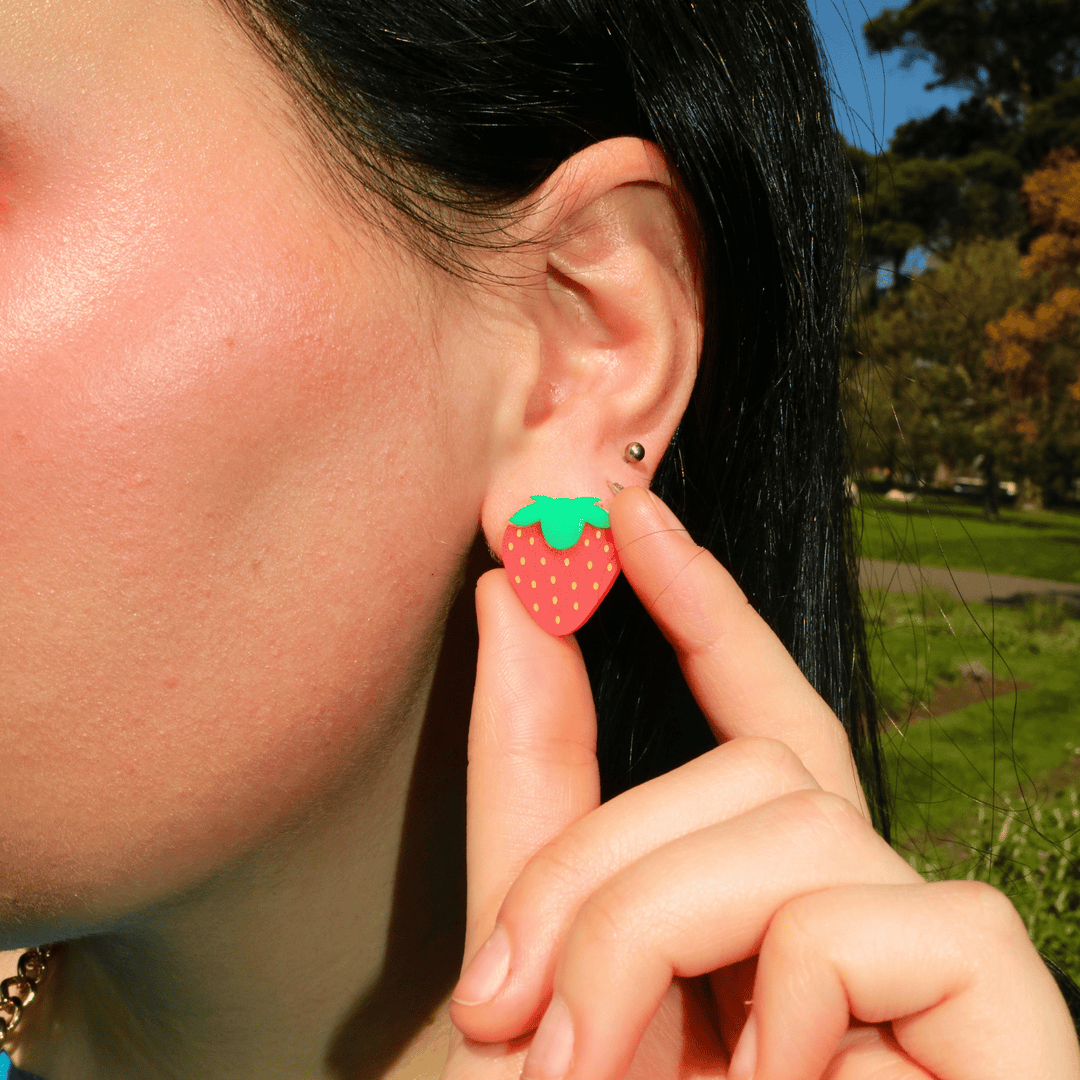 close up shot of model wearing haus of dizzy strawberry studs with model holding stud between pointer finger and thumb. the studs use a frosted red acrylic with yellow painted seeds and a green acrylic top