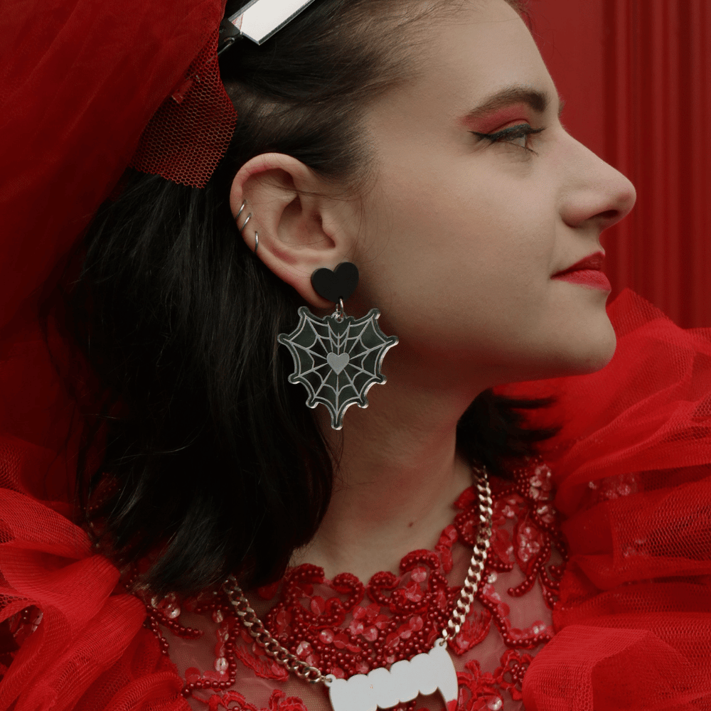 close up image of model wearing haus of dizzy web of lies silver mirror acrylic earrings. the earrings are a spider web in the shape of a heart with the etched acrylic resembling a spider web