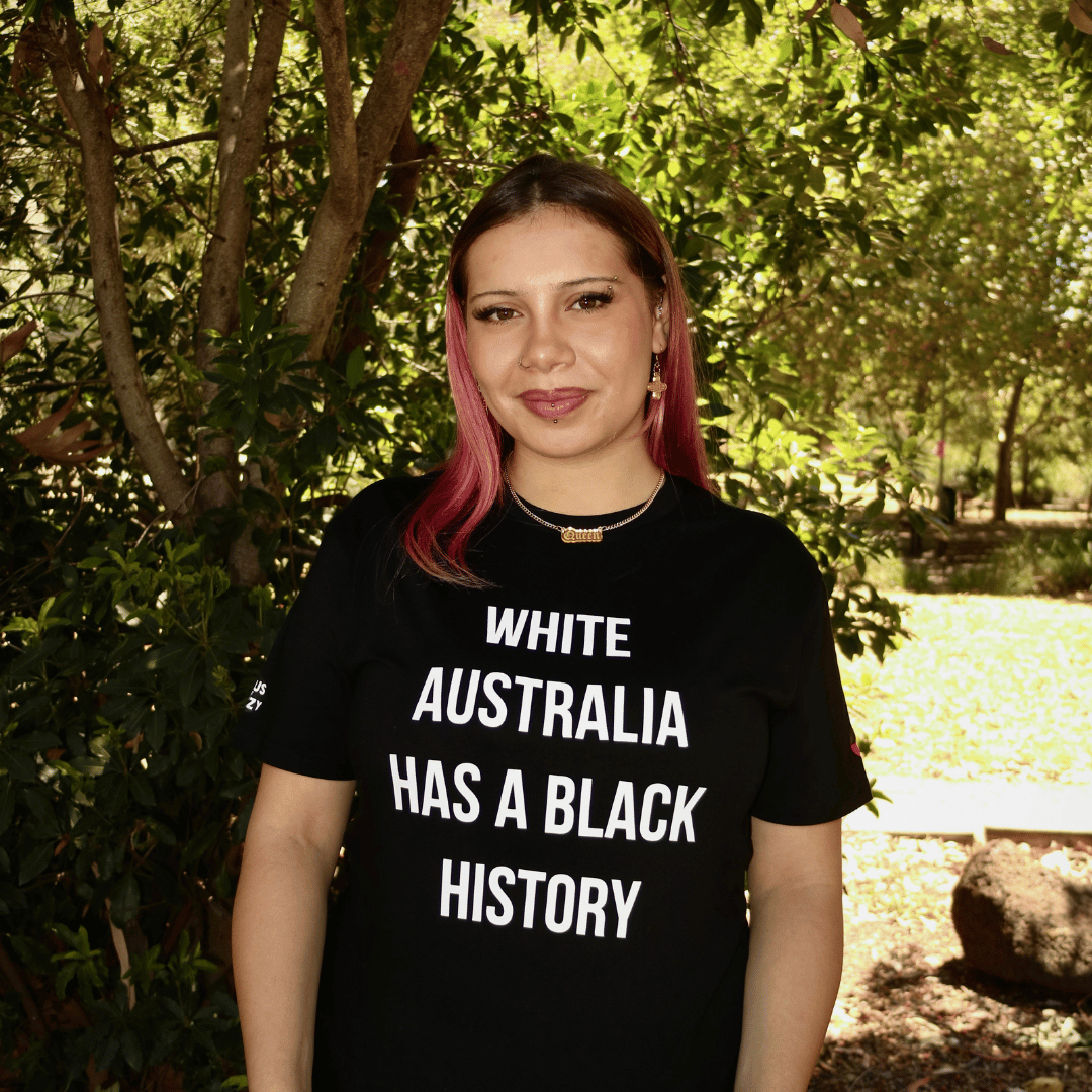Person wearing haus of dizzy black t-shirt with 'White Australia Has a Black History' text in a park setting.