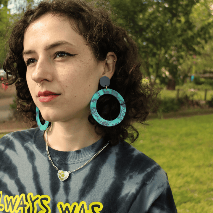 model wearing haus of dizzy wiradjrui yinaa earth tones hoop earrings in dusty teal colour, outdoors with greenery in the background
